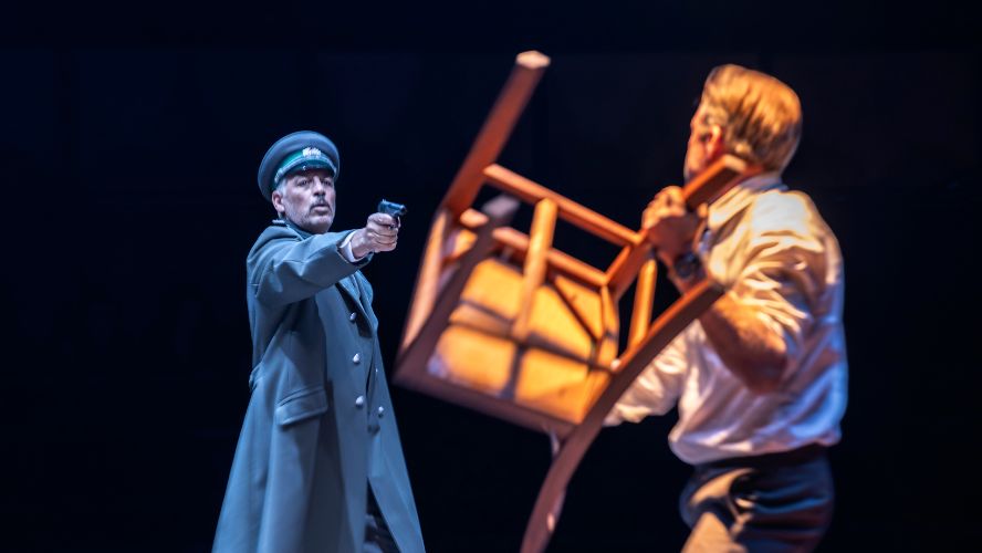 A man in a dark uniform is holding a gun towards another man, who is holding a chair as if to throw it