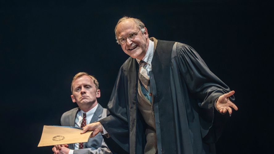 John Ramm plays Karden wearing a black gown and holding a manila folder