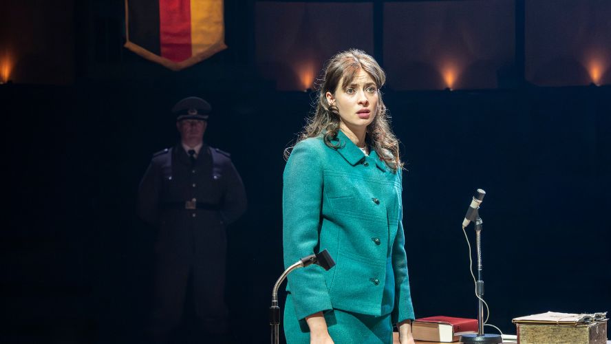 Agnes O'Casey plays Liz Gold, wearing a turquoise suit standing next to a microphone