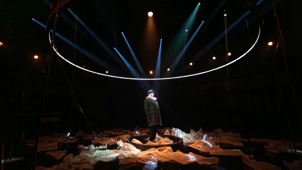 Phelim McDermott stands in the centre of the stage, surrounded by sheet music on the floor, wearing a long coast and bowler hat