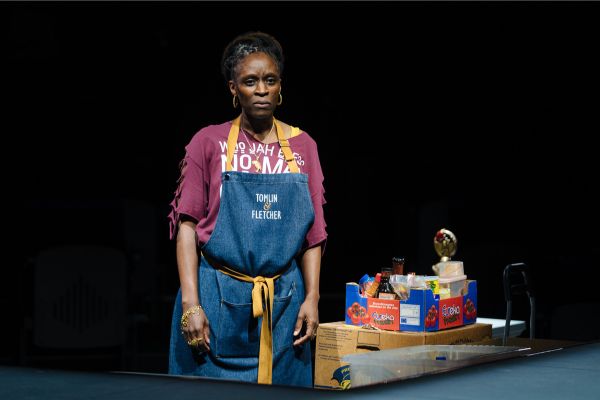 Denise (played by Sharon Duncan Brewster) wears an apron that says 'Tomlin & Fletcher'