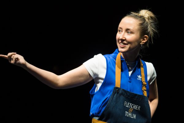 Carly (played by Erin Doherty) is pointing, wearing an apron that says 'Fletcher & Tomlin'