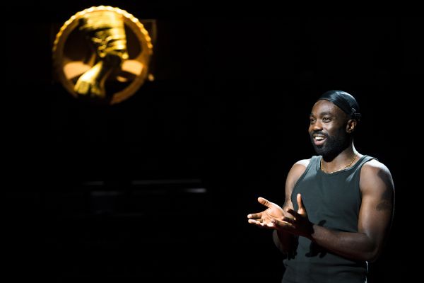 Delroy (played by Paapa Essiedu) is in the foreground with a golden statue of Nefertiti in the background