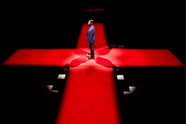 Delroy (played by Paapa Essiedu) stands in the middle of a red cross