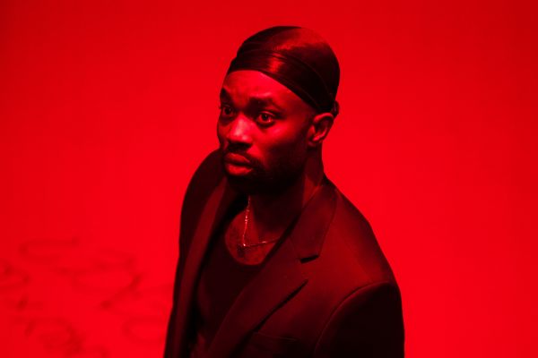 Delroy (played by Paapa Essiedu) wears a suit on a red lit stage