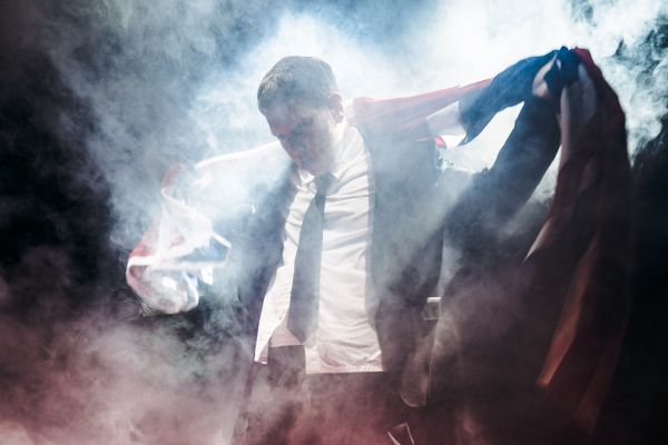 Michael (played by Thomas Coombes) wears a suit, holding the Union Jack flag around his shoulders, surrounded by haze