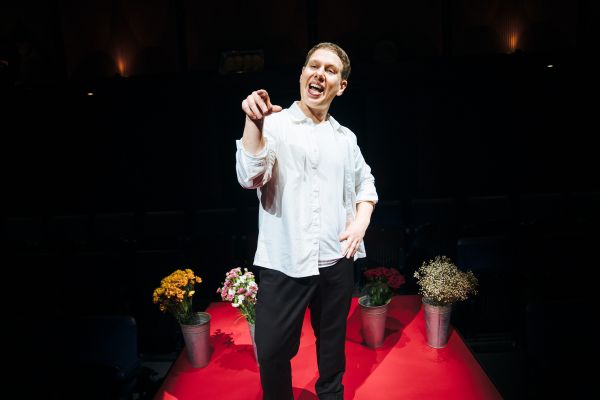 Michael (played by Thomas Coombes) points towards the audience with four buckets of flowers on the floor behind him